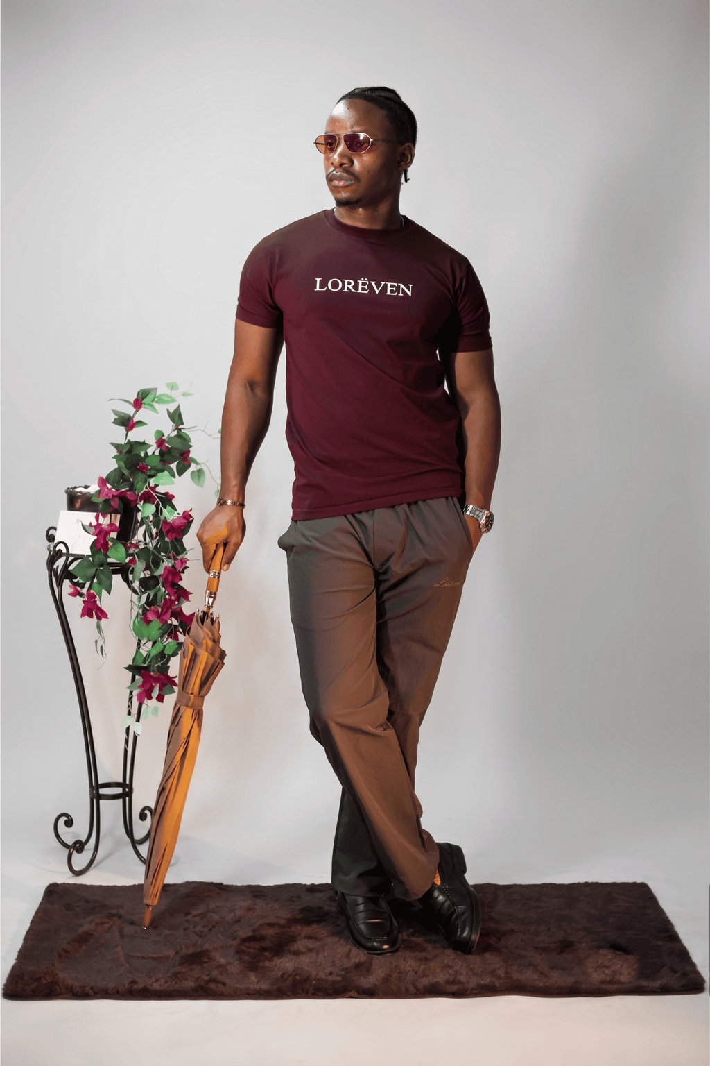 LORËVEN Drift Stretch Trouser in Anchor Charcoal — man in a burgundy LORËVEN t-shirt holding a wooden umbrella beside a floral stand against a light studio background