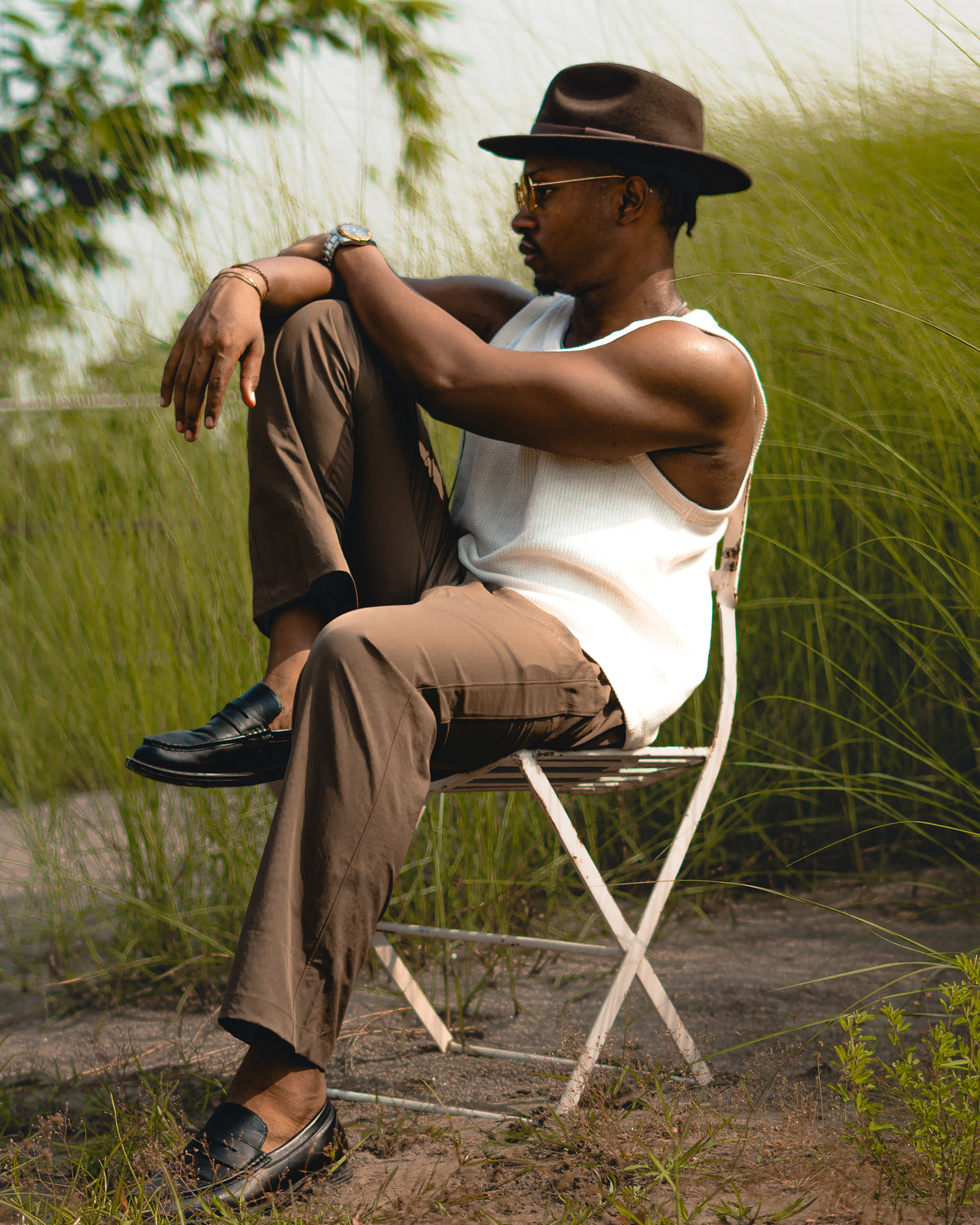 LORËVEN Drift Stretch Trouser in Mouse Brown — man in a brown fedora and white tank top sitting on a white metal folding chair outdoors in a grassy field