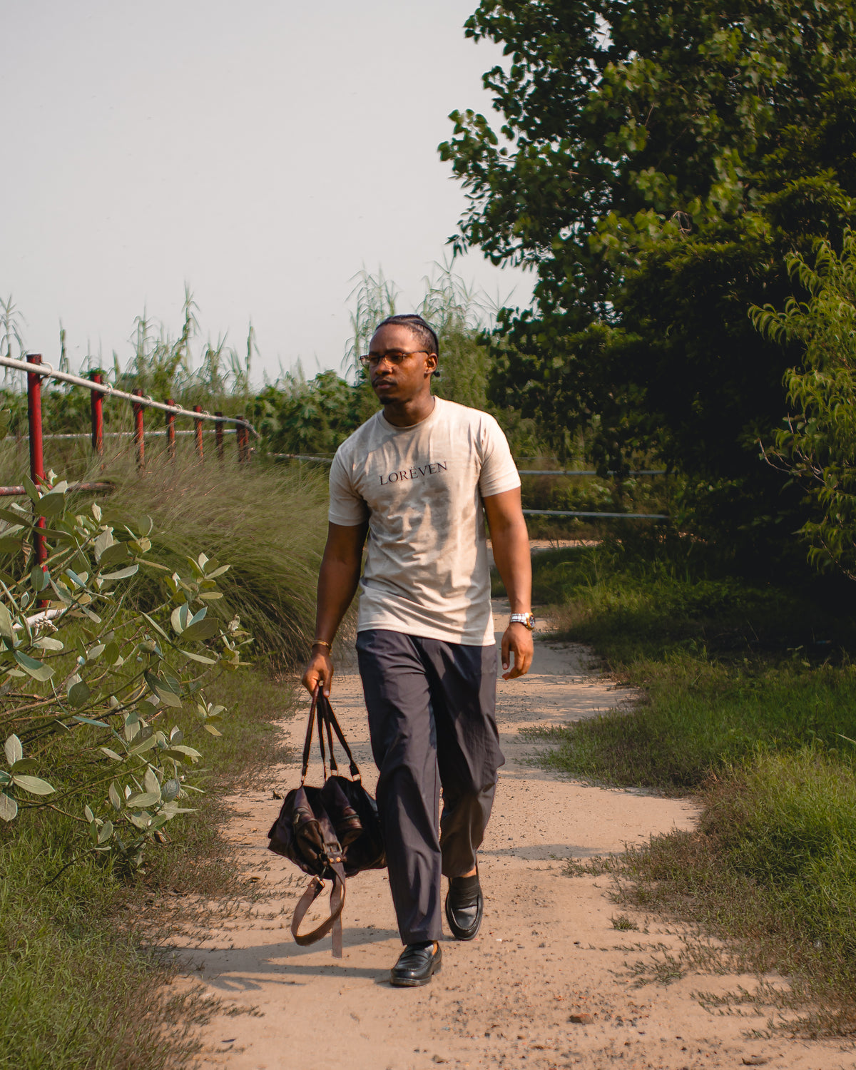 LORËVEN Drift Stretch Trouser in Slate Grey — man walking on a dirt path outdoors wearing a white LORËVEN t-shirt and carrying a black duffel bag