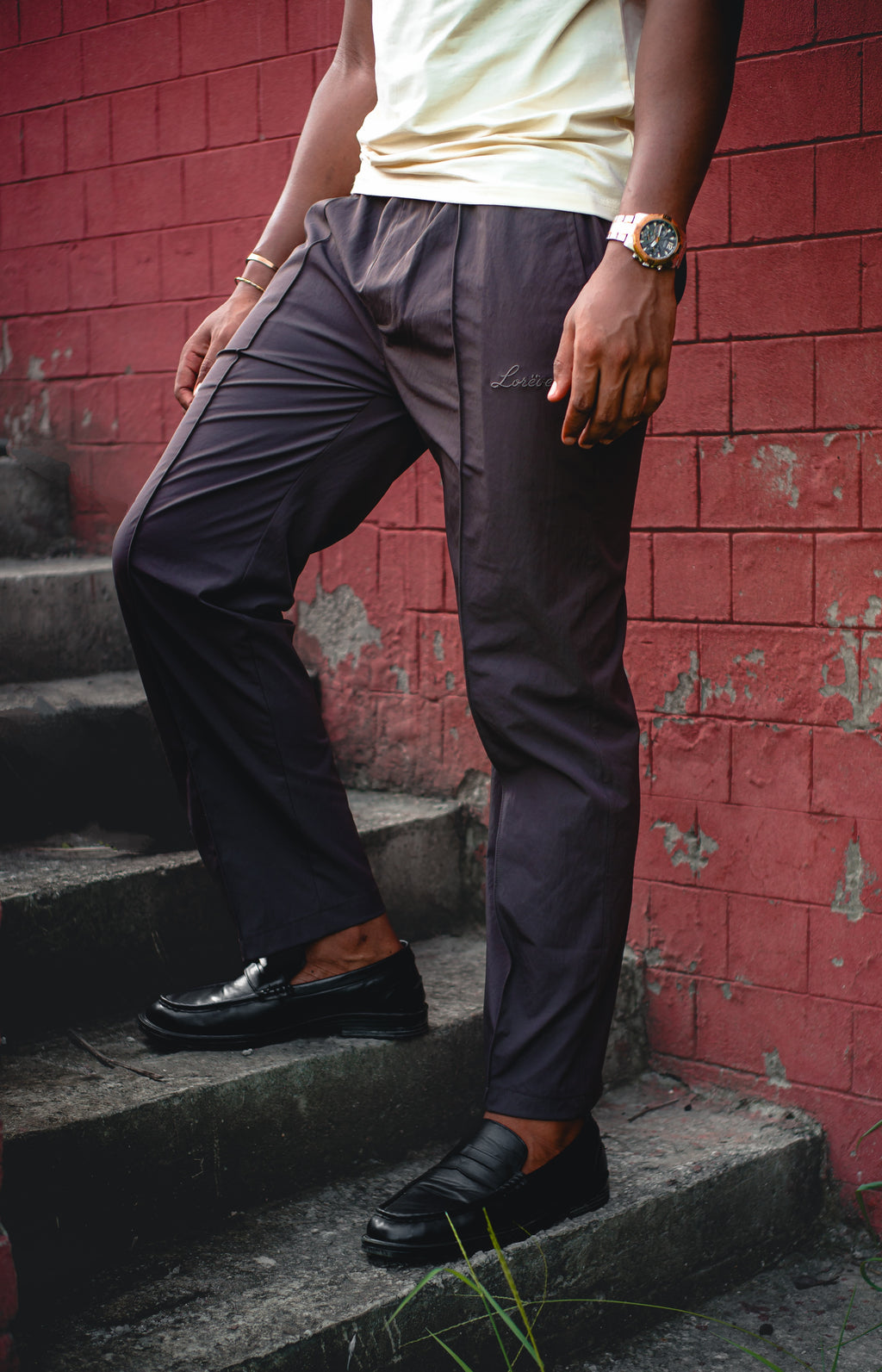 LORËVEN Fold Stretch Trouser in Anchor Charcoal — close-up of a man in a pale yellow t-shirt and black loafers descending concrete steps against a red brick wall, wearing tailored charcoal trousers with a tonal embroidered logo