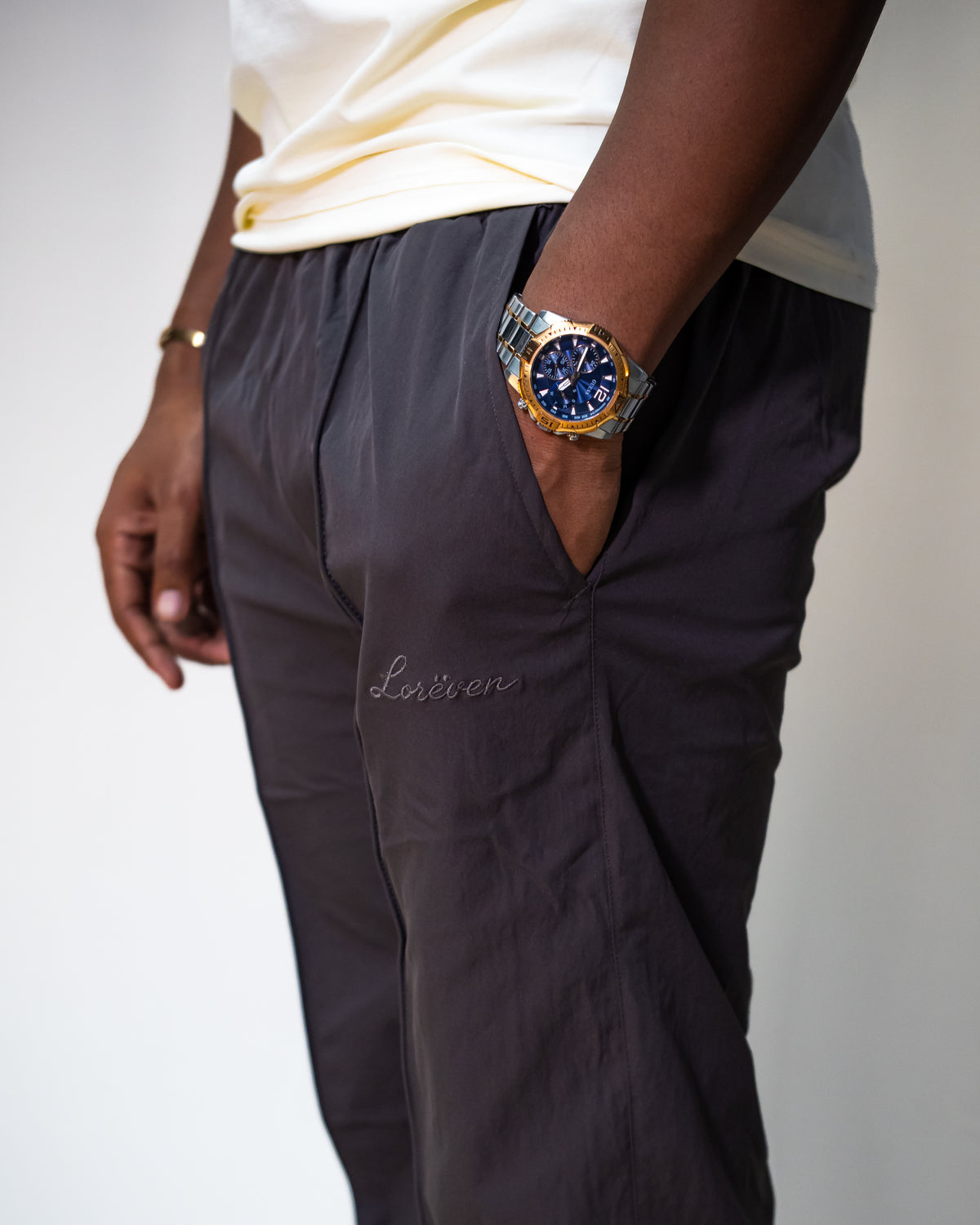 LORËVEN Fold Stretch Trouser in Anchor Charcoal — close-up of the drawstring waist and hip pocket with tonal embroidered script logo, paired with a cream t-shirt