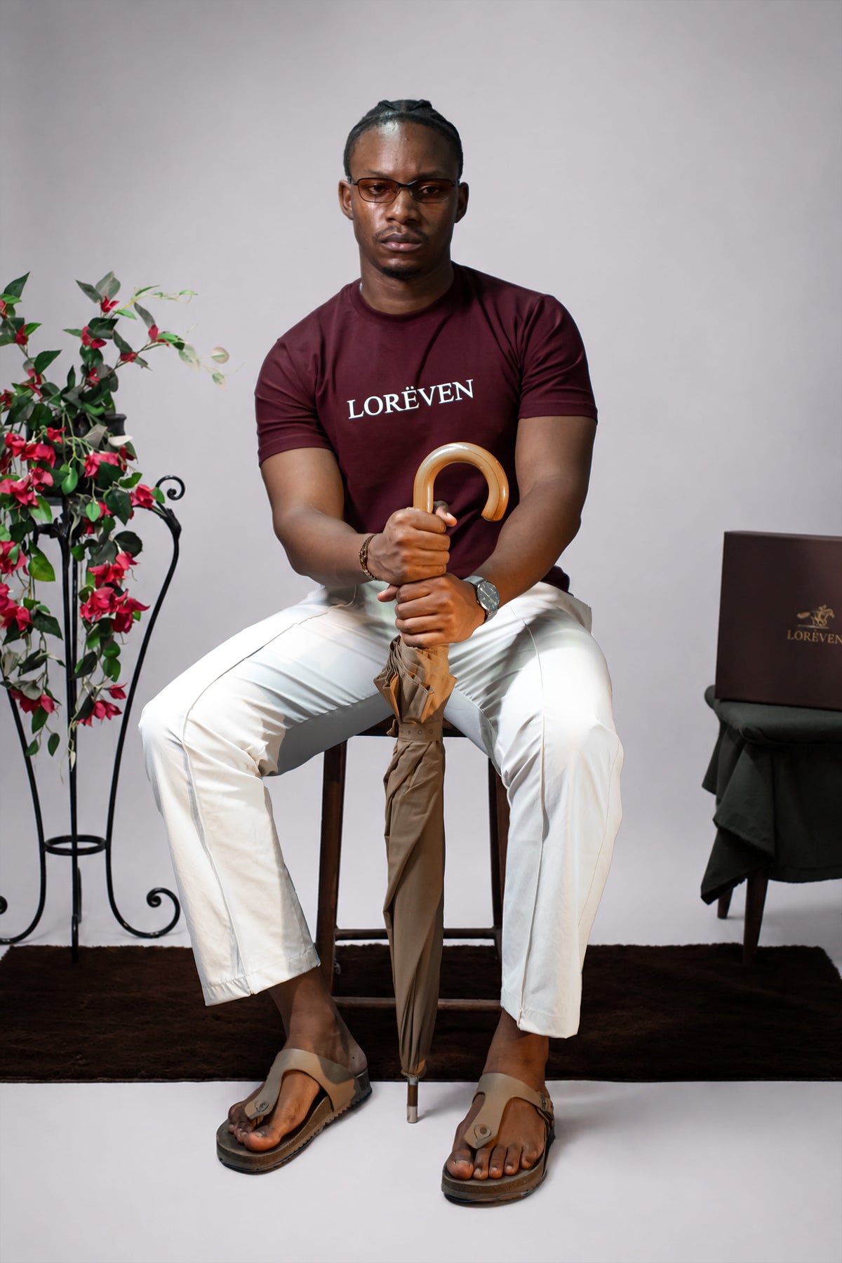 LORËVEN Fold Stretch Trouser in Chalk White — man in a maroon LORËVEN t-shirt sitting on a wooden stool holding a closed umbrella, against a neutral grey background