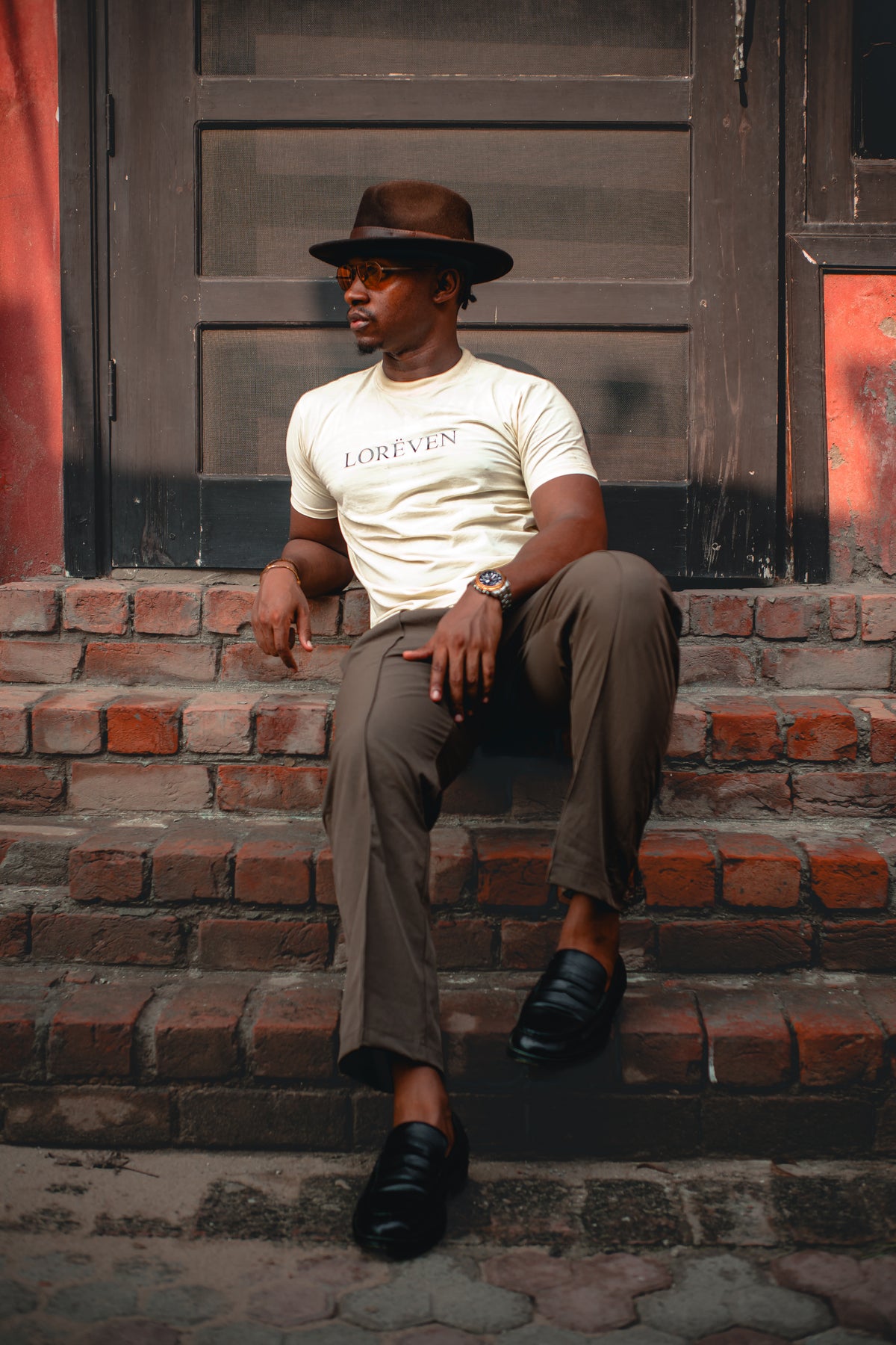 LORËVEN Fold Stretch Trouser in Mouse Brown — man wearing a brown fedora, cream LORËVEN t-shirt, and black loafers, sitting on red brick steps in front of a dark wooden door