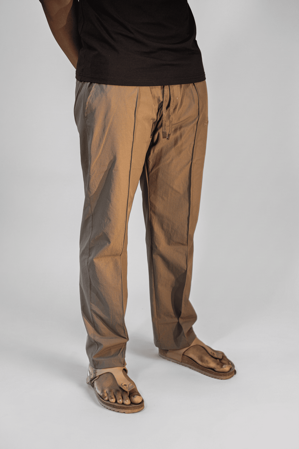 LORËVEN Fold Stretch Trouser in Mouse Brown — close-up of the tailored trouser leg with drawstring waist and side pocket, paired with a black t-shirt and brown sandals against a plain studio background