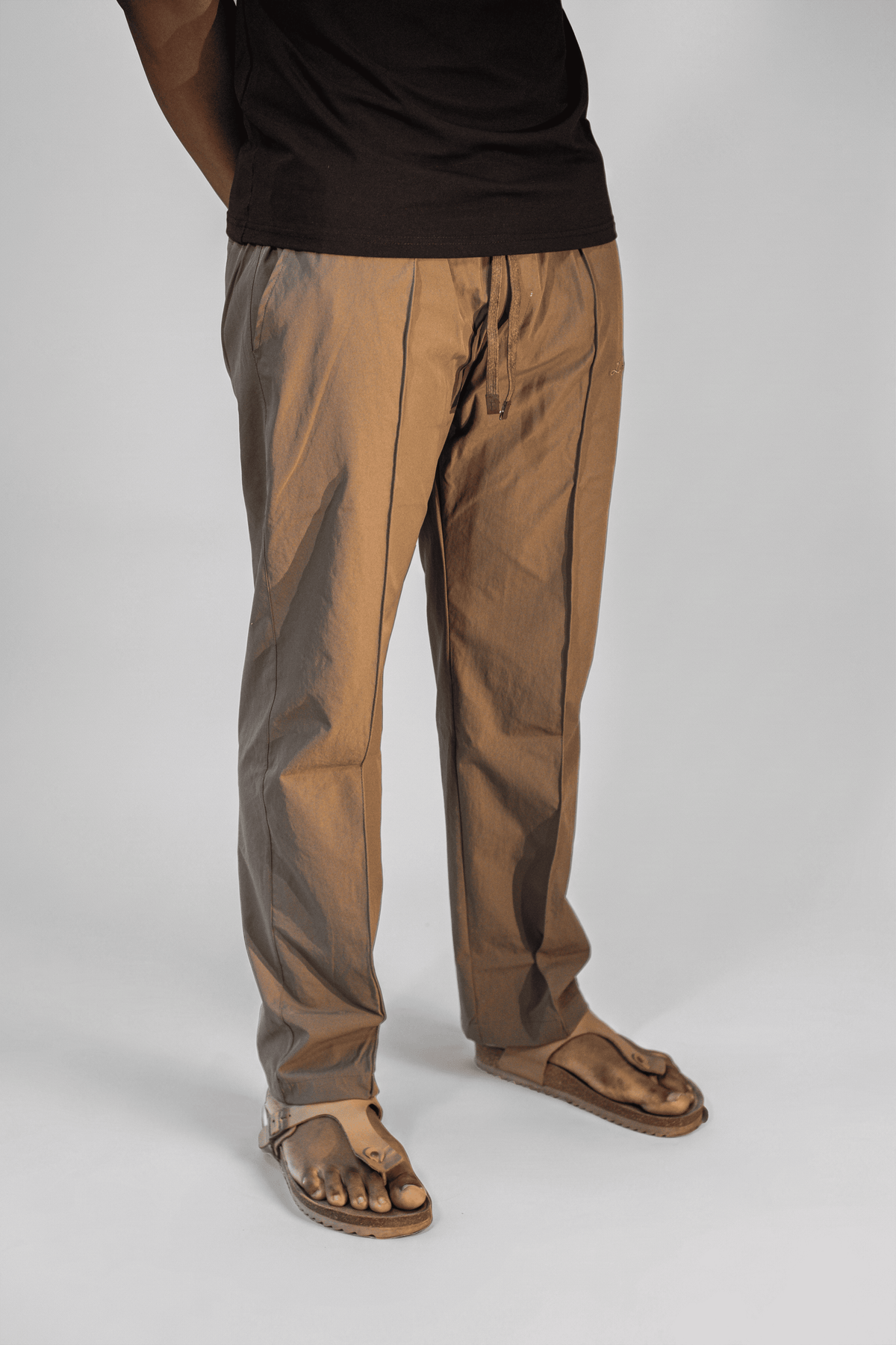 LORËVEN Fold Stretch Trouser in Mouse Brown — close-up of the tailored trouser leg with drawstring waist and side pocket, paired with a black t-shirt and brown sandals against a plain studio background