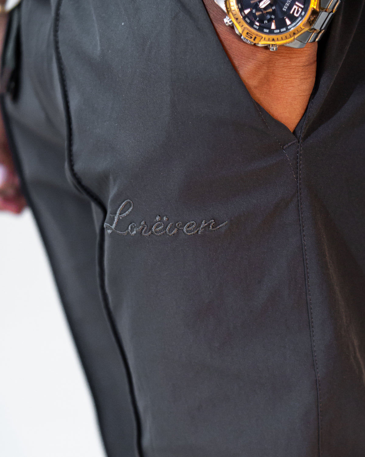 LORËVEN Fold Stretch Trouser in Slate Grey — macro close-up of the tonal embroidered "Lorëven" script logo on the pocket, with a gold and black watch visible