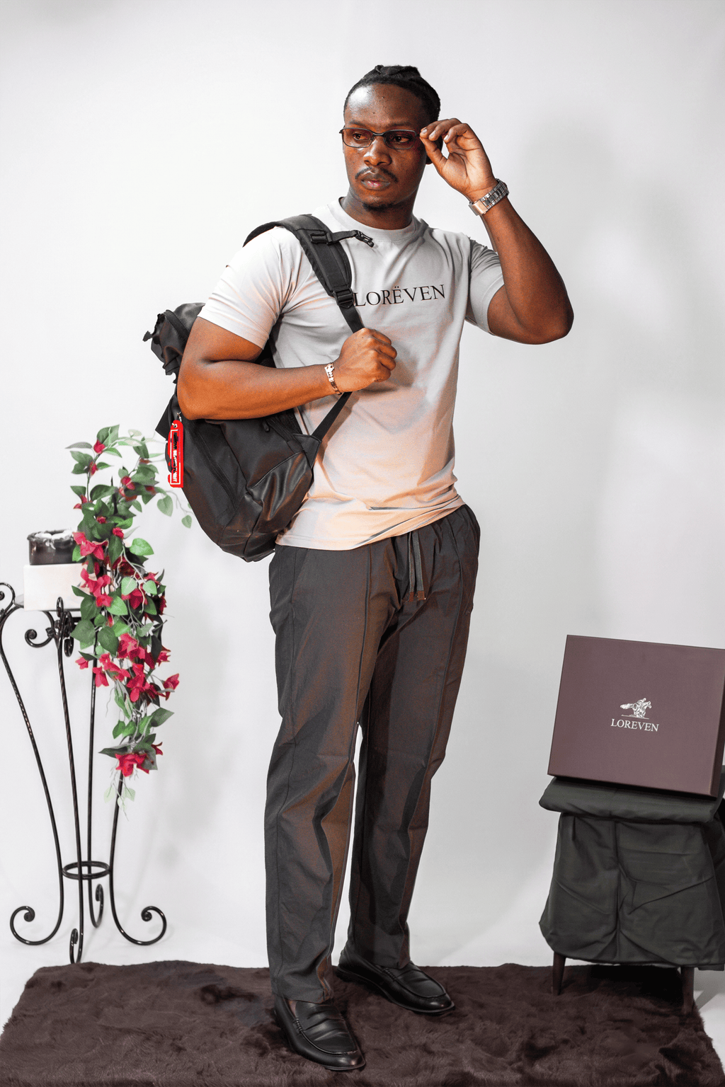 LORËVEN Fold Stretch Trouser in Slate Grey — man wearing a light beige LORËVEN t-shirt and black shoes, adjusting his glasses with a black backpack on his shoulder, standing in a studio setting with a decorative plant and a branded box