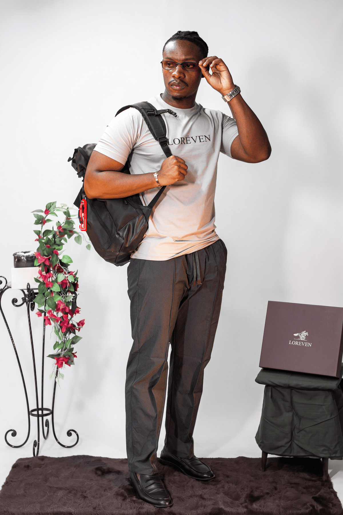 LORËVEN Fold Stretch Trouser in Slate Grey — man wearing a light beige LORËVEN t-shirt and black shoes, adjusting his glasses with a black backpack on his shoulder, standing in a studio setting with a decorative plant and a branded box