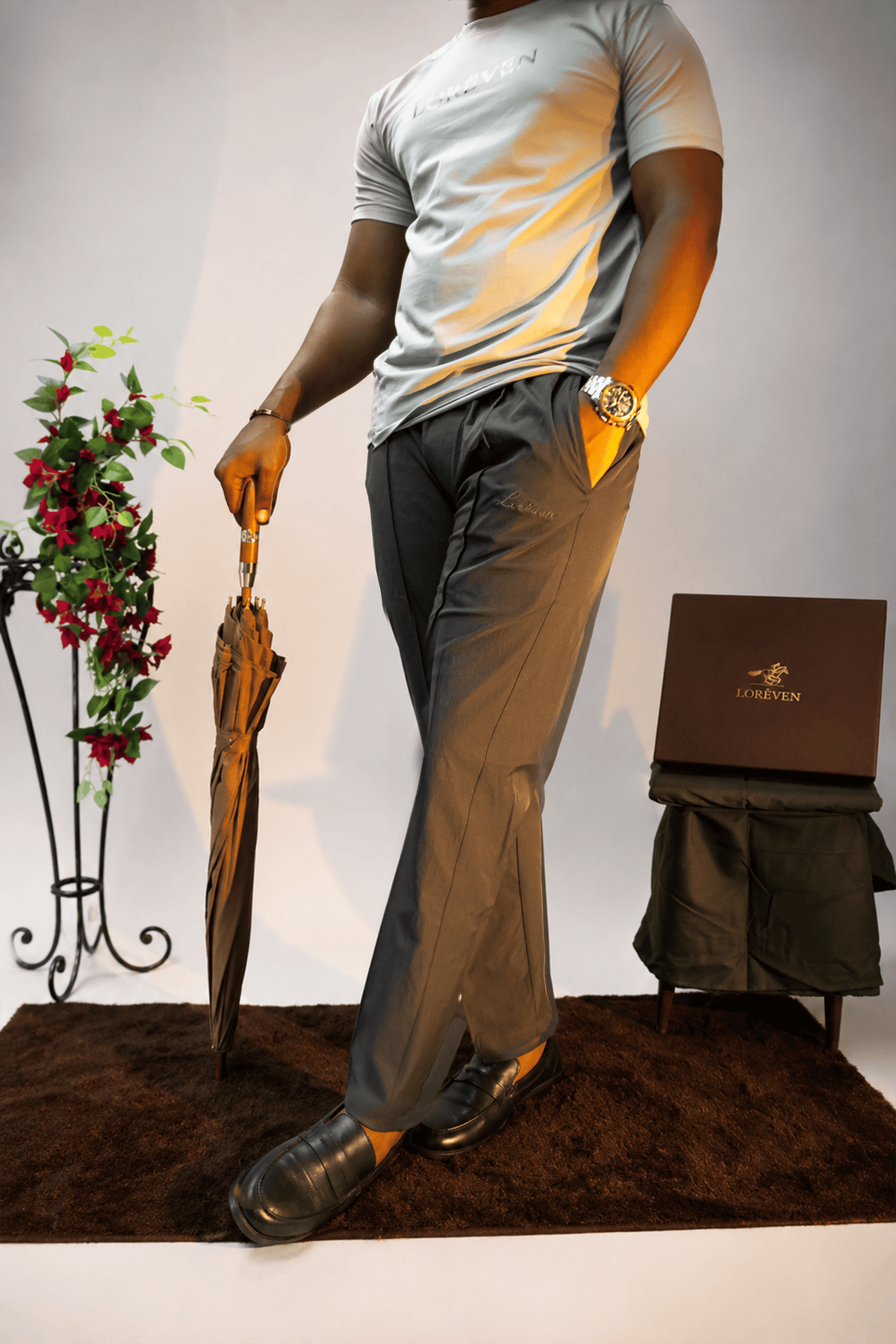 LORËVEN Fold Stretch Trouser in Slate Grey — man in a light grey LORËVEN t-shirt holding a closed umbrella, standing on a brown rug with a potted plant and a LORËVEN branded box on a stool against a white studio background