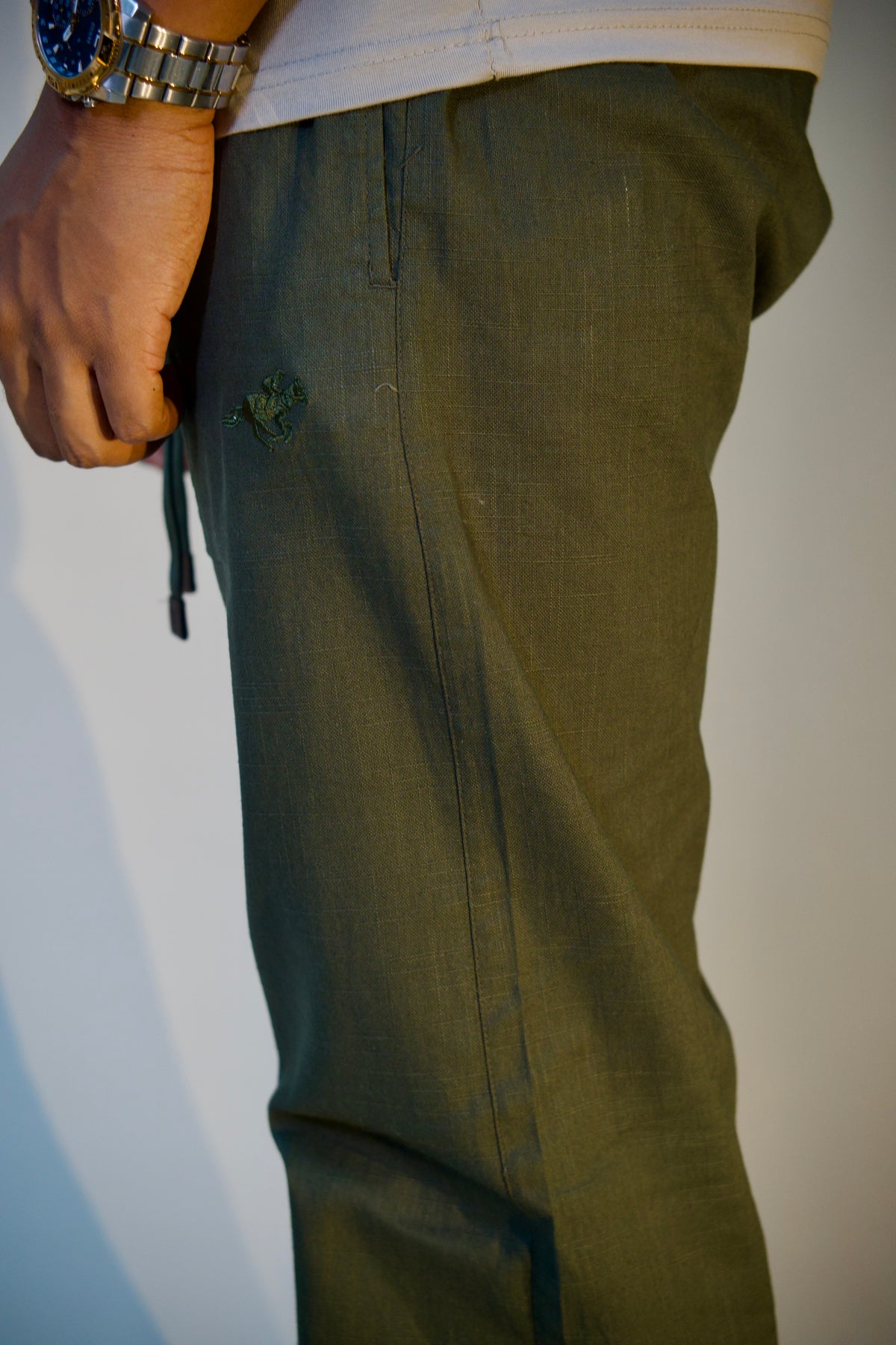 Close-up of a person wearing olive green pants, showing the side pocket with a small embroidered logo and a drawstring, against a plain light background.