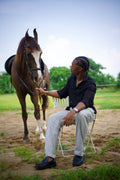 LORËVEN Linen Cotton Trouser in Silver Moon grey — man in a black shirt seated on a folding chair, holding the reins of a horse in an outdoor paddock