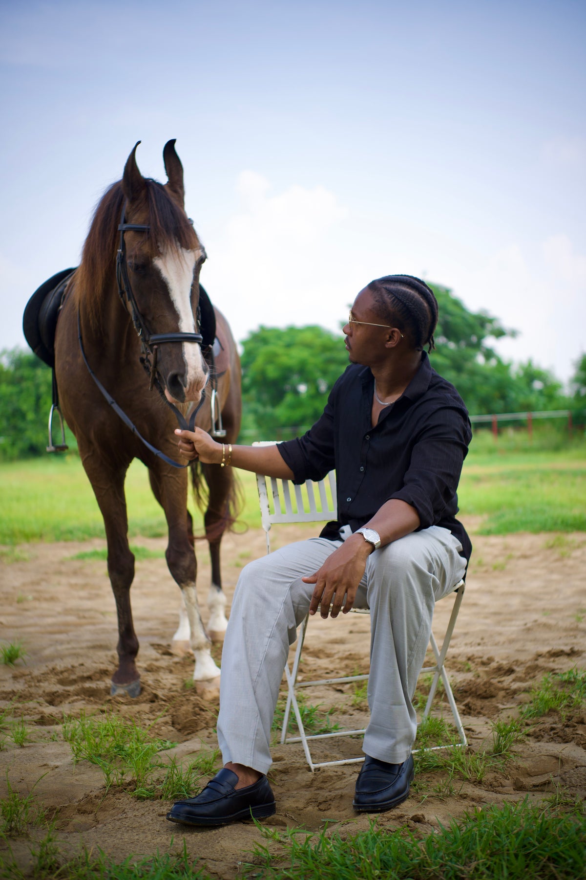 LORËVEN Linen Cotton Trouser in Silver Moon grey — man in a black shirt seated on a folding chair, holding the reins of a horse in an outdoor paddock