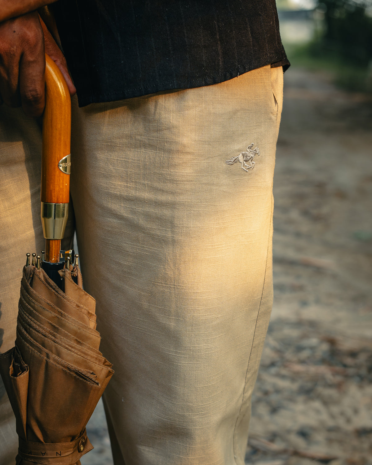 Close-up of LORËVEN linen trousers in beige with tonal horse embroidery, worn with a wooden-handled umbrella