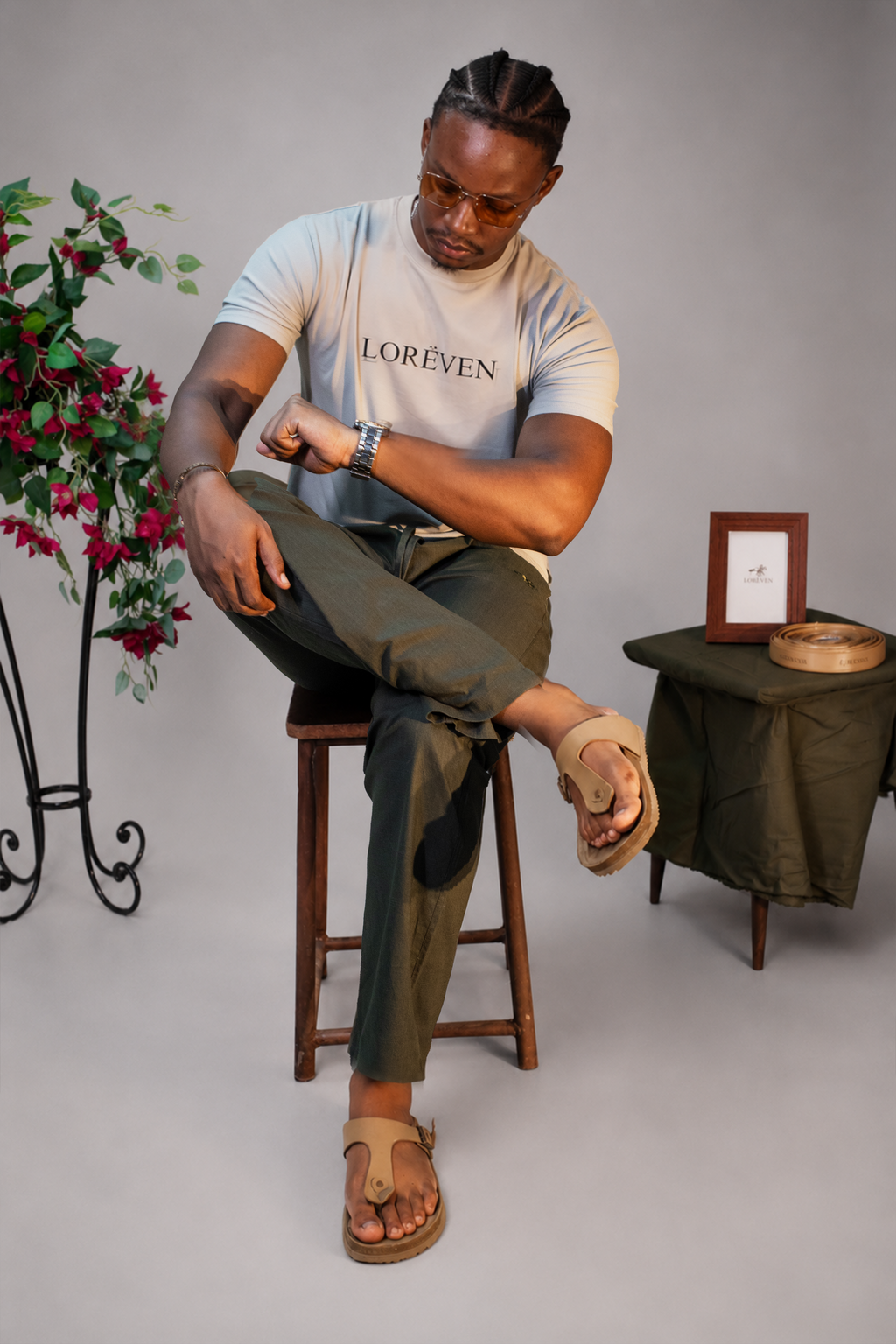 LORËVEN Linen Cotton Trouser in Deep Olive — man in a light beige LORËVEN t-shirt and tan leather sandals seated on a wooden stool in a studio setting with a floral arrangement
