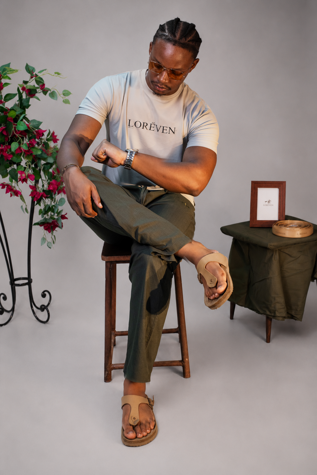 LORËVEN Linen Cotton Trouser in Deep Olive — man in a light beige LORËVEN t-shirt and tan leather sandals seated on a wooden stool in a studio setting with a floral arrangement