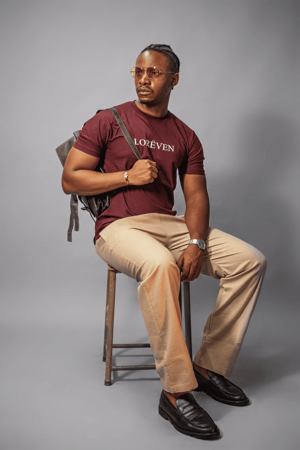 LORËVEN Linen Cotton Trouser in Moccasin Tan — man in a burgundy LORËVEN t-shirt and black loafers seated on a wooden stool against a grey studio background