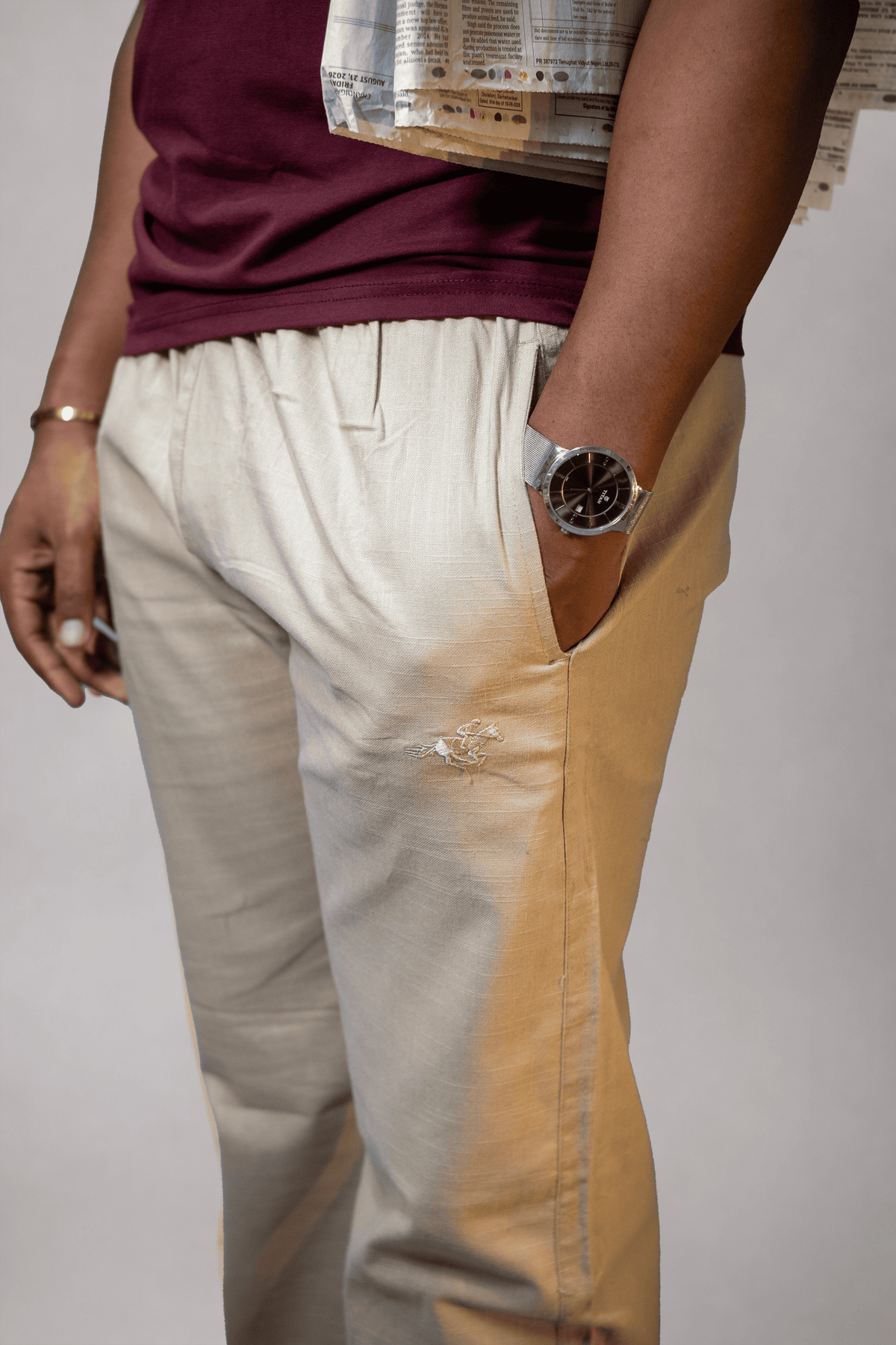 LORËVEN Linen Cotton Trouser in Moccasin Tan — close-up of the trouser leg and front pocket with tonal embroidered detail, paired with a maroon top and silver watch