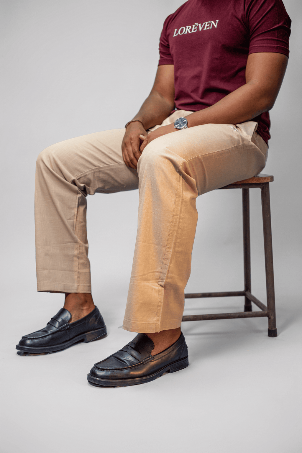 LORËVEN Linen Cotton Trouser in Moccasin Tan — close-up of a man seated on a wooden stool, wearing a burgundy LORËVEN t-shirt and black leather loafers against a light grey studio background