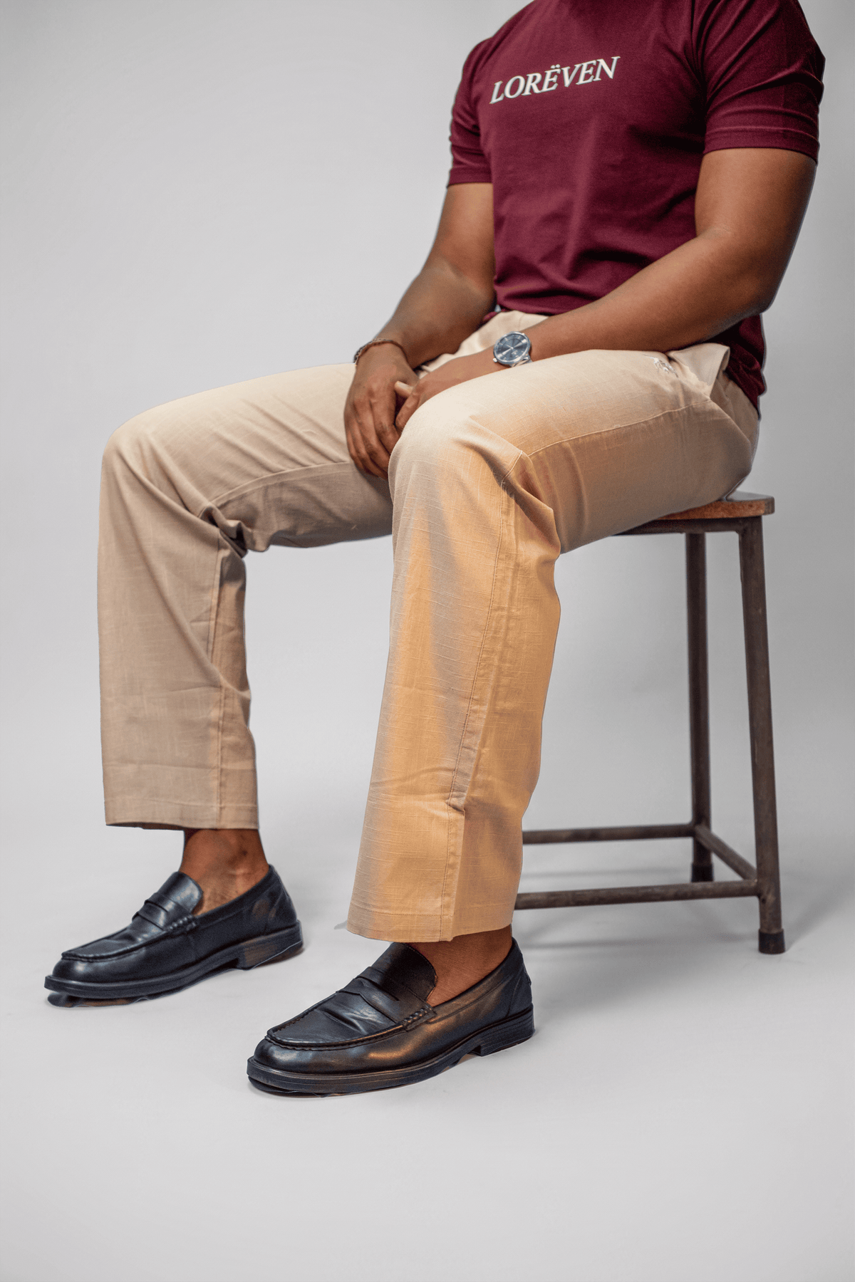 LORËVEN Linen Cotton Trouser in Moccasin Tan — close-up of a man seated on a wooden stool, wearing a burgundy LORËVEN t-shirt and black leather loafers against a light grey studio background