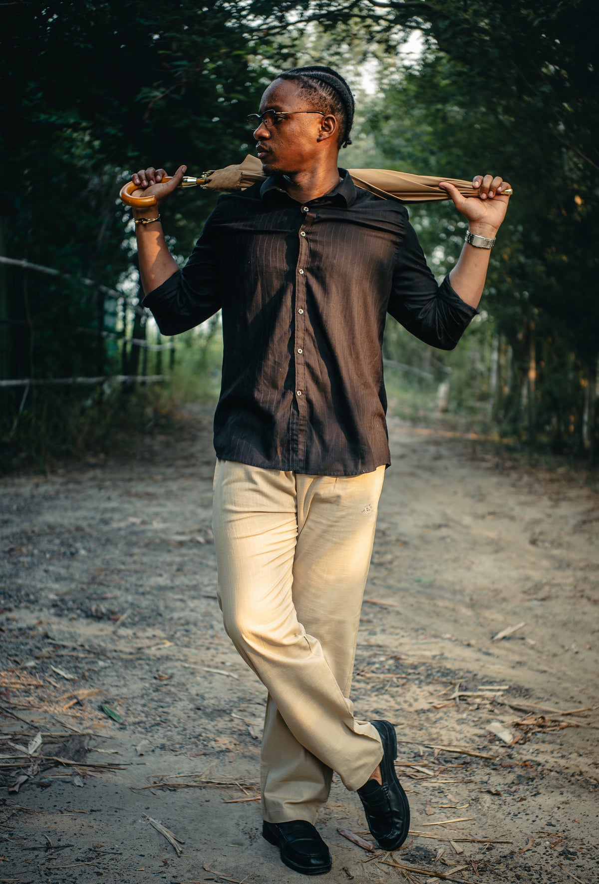 LORËVEN Linen Cotton Trouser in Moccasin Tan — man in a black button-up shirt walking along a dirt path outdoors, carrying a tan umbrella over his shoulder