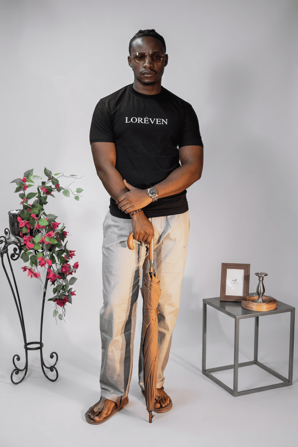 LORËVEN Linen Cotton Trouser in Silver Moon grey — man in a black LORËVEN t-shirt and brown sandals standing in a studio with a rolled umbrella and floral arrangement