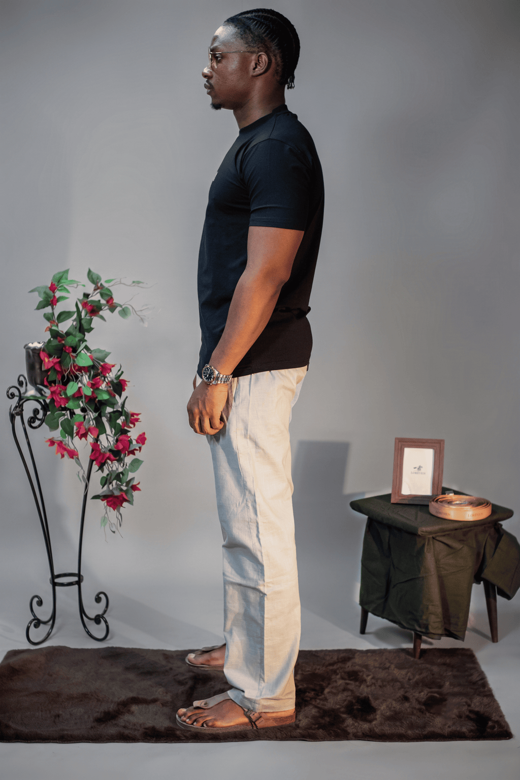 LORËVEN Linen Cotton Trouser in Silver Moon grey — man in a black t-shirt shown in profile, standing on a dark rug in a studio setting with a floral arrangement and side table
