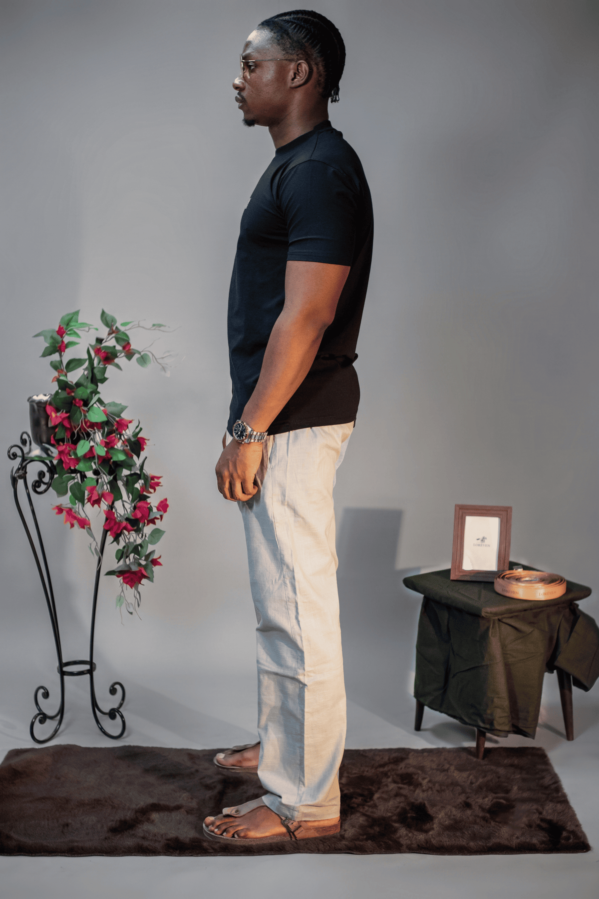 LORËVEN Linen Cotton Trouser in Silver Moon grey — man in a black t-shirt shown in profile, standing on a dark rug in a studio setting with a floral arrangement and side table
