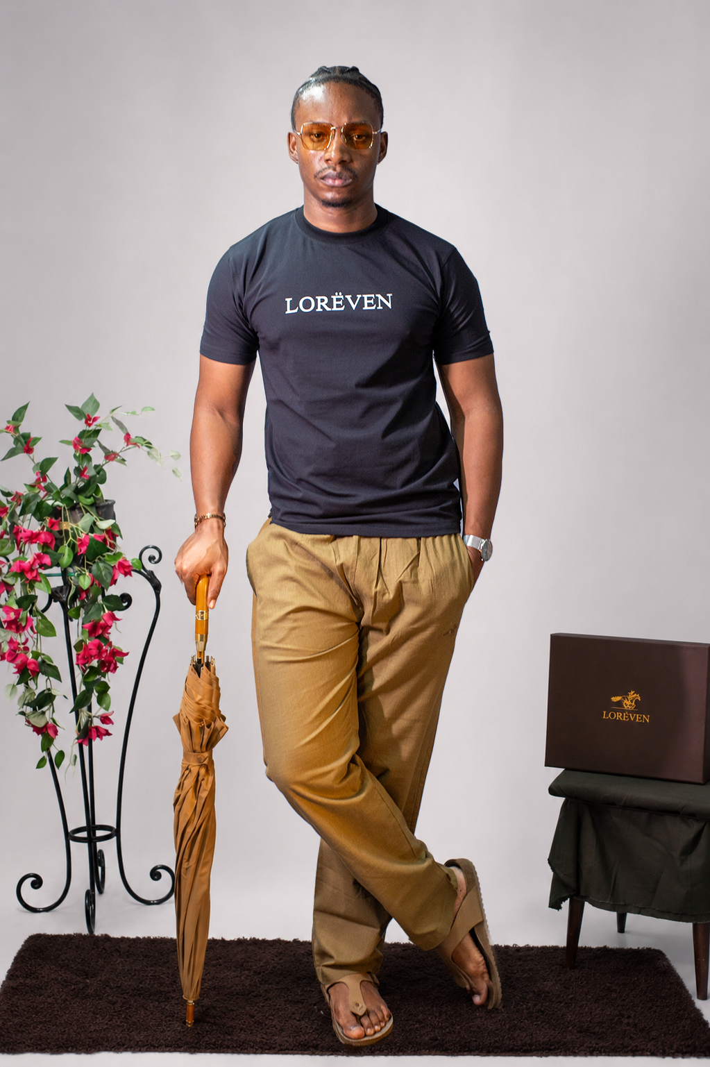 LORËVEN Linen Cotton Trouser in Tobacco Brown — man in a black LORËVEN t-shirt holding a brown umbrella, standing in a studio setting with a floral arrangement and a LORËVEN box