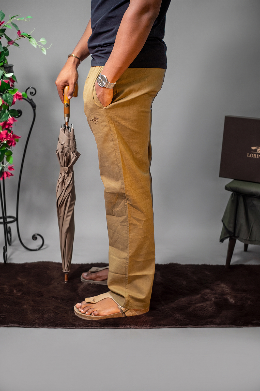 LORËVEN Linen Cotton Trouser in Tobacco Brown — side profile of a man holding a brown umbrella, standing on a dark rug in a studio setting with a floral stand and a LORËVEN box