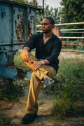 LORËVEN Linen Cotton Trouser in Tobacco Brown — man in a black button-down shirt seated on a weathered blue metal container outdoors