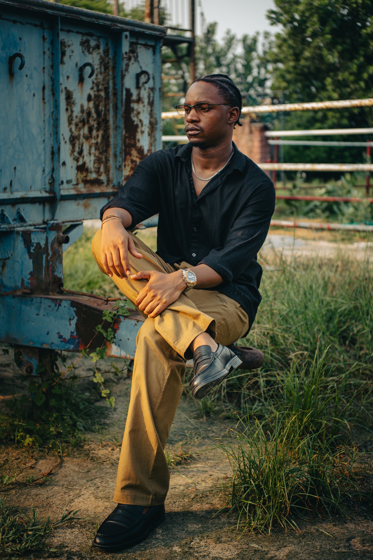 LORËVEN Linen Cotton Trouser in Tobacco Brown — man in a black button-down shirt seated on a weathered blue metal container outdoors