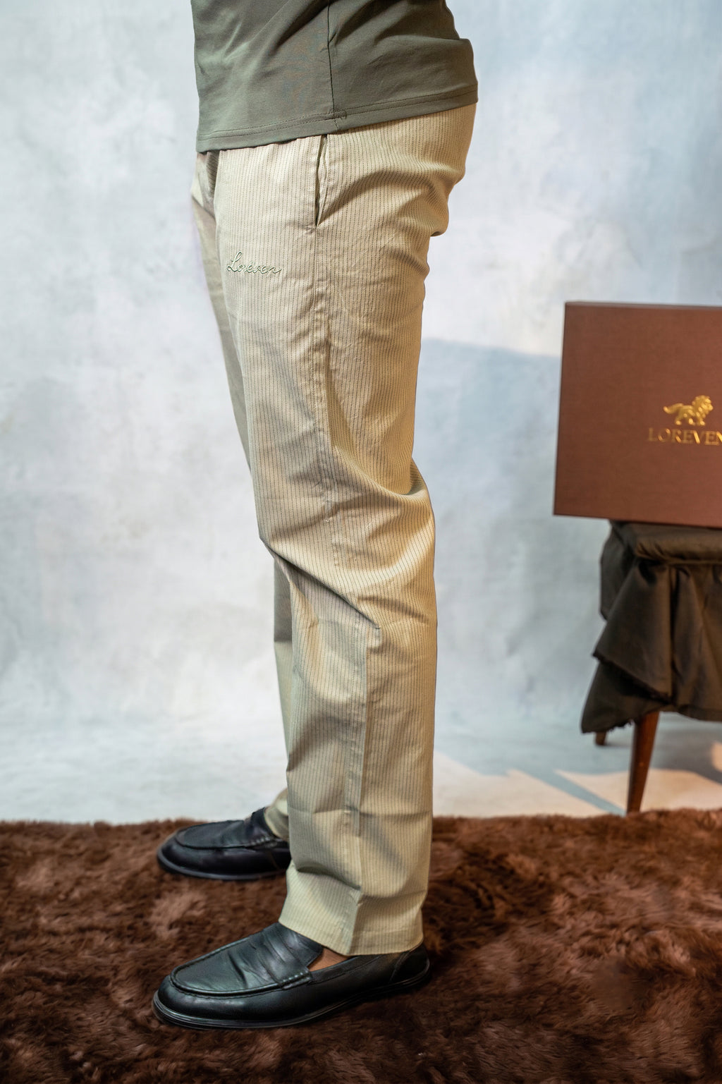 LORËVEN Striped Linen Trouser in Lichen Sage — close-up of the trouser leg with embroidered logo, paired with black loafers and an olive green shirt, standing on a faux fur rug