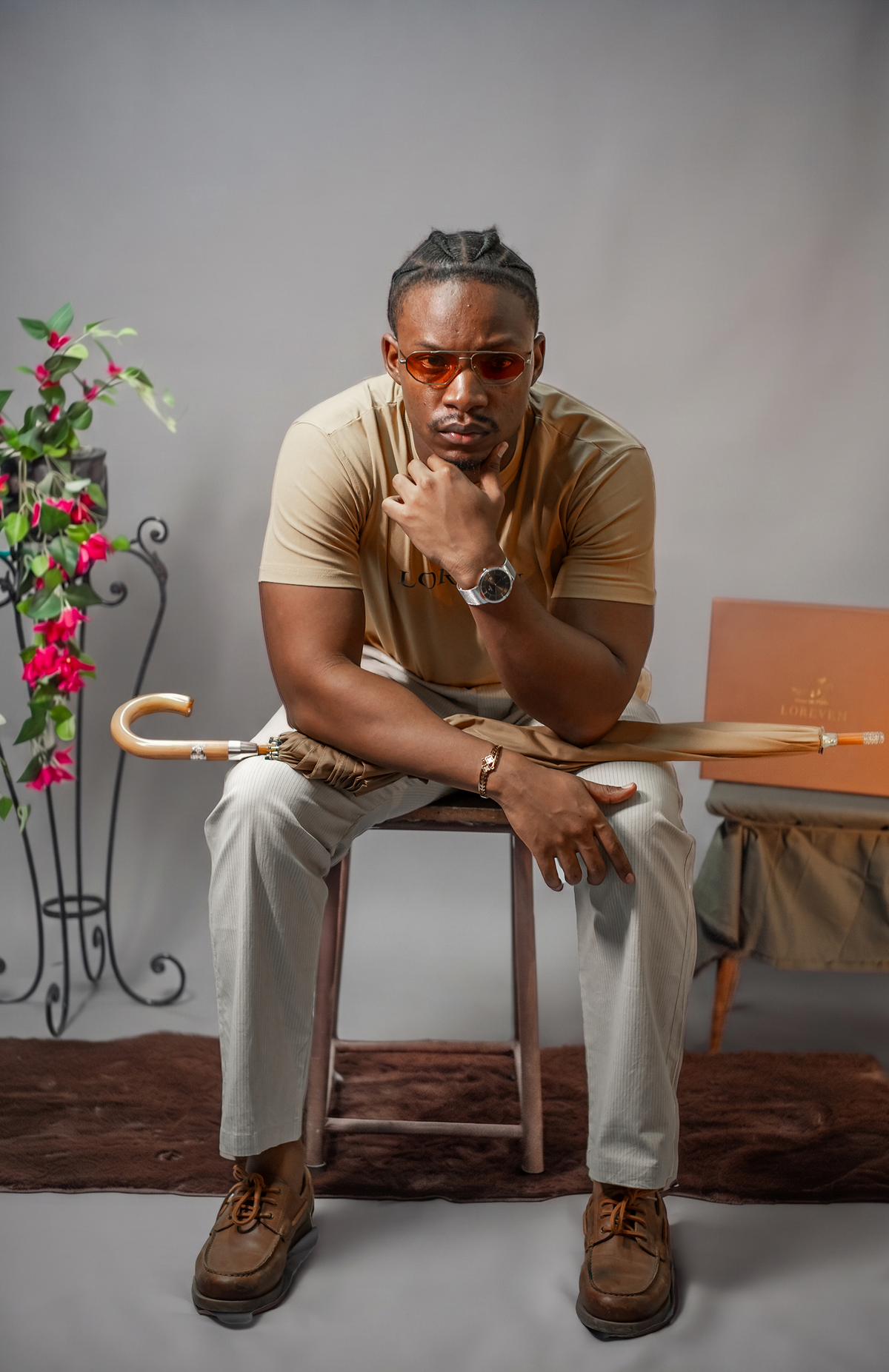 LORËVEN Striped Linen Trouser in Dune Beige — man in a beige t-shirt and tinted sunglasses seated on a wooden stool, holding a wooden-handled umbrella in a studio setting with a floral arrangement