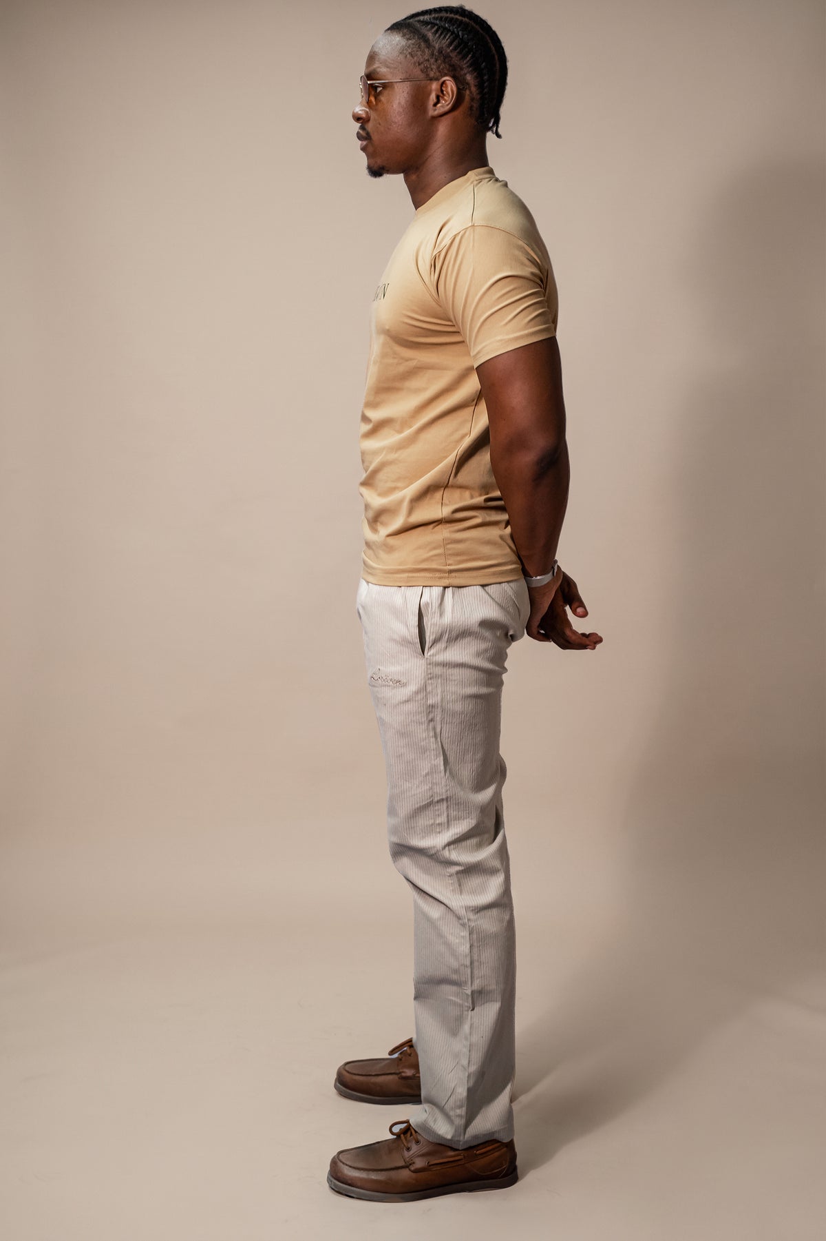 LORËVEN Striped Linen Trouser in Dune Beige — man in profile wearing a tan crew-neck t-shirt and brown leather loafers, standing against a beige studio background