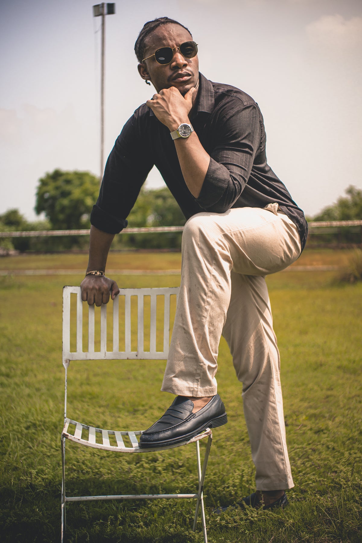 LORËVEN Striped Linen Trouser in Dune Beige — man in sunglasses and a black shirt seated on a white metal chair in an outdoor grassy field