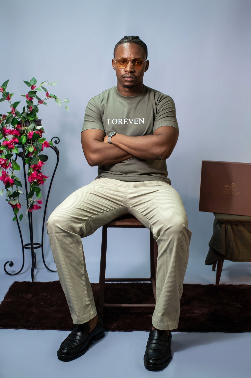 LORËVEN Striped Linen Trouser in Lichen Sage — man in a sage green LORËVEN t-shirt and tinted sunglasses seated on a wooden stool against a light blue-grey background