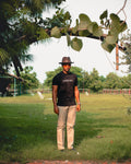LORËVEN Striped Linen Trouser in Lichen Sage — man in a black LORËVEN t-shirt and brown fedora hat standing in a grassy outdoor park under a tree branch
