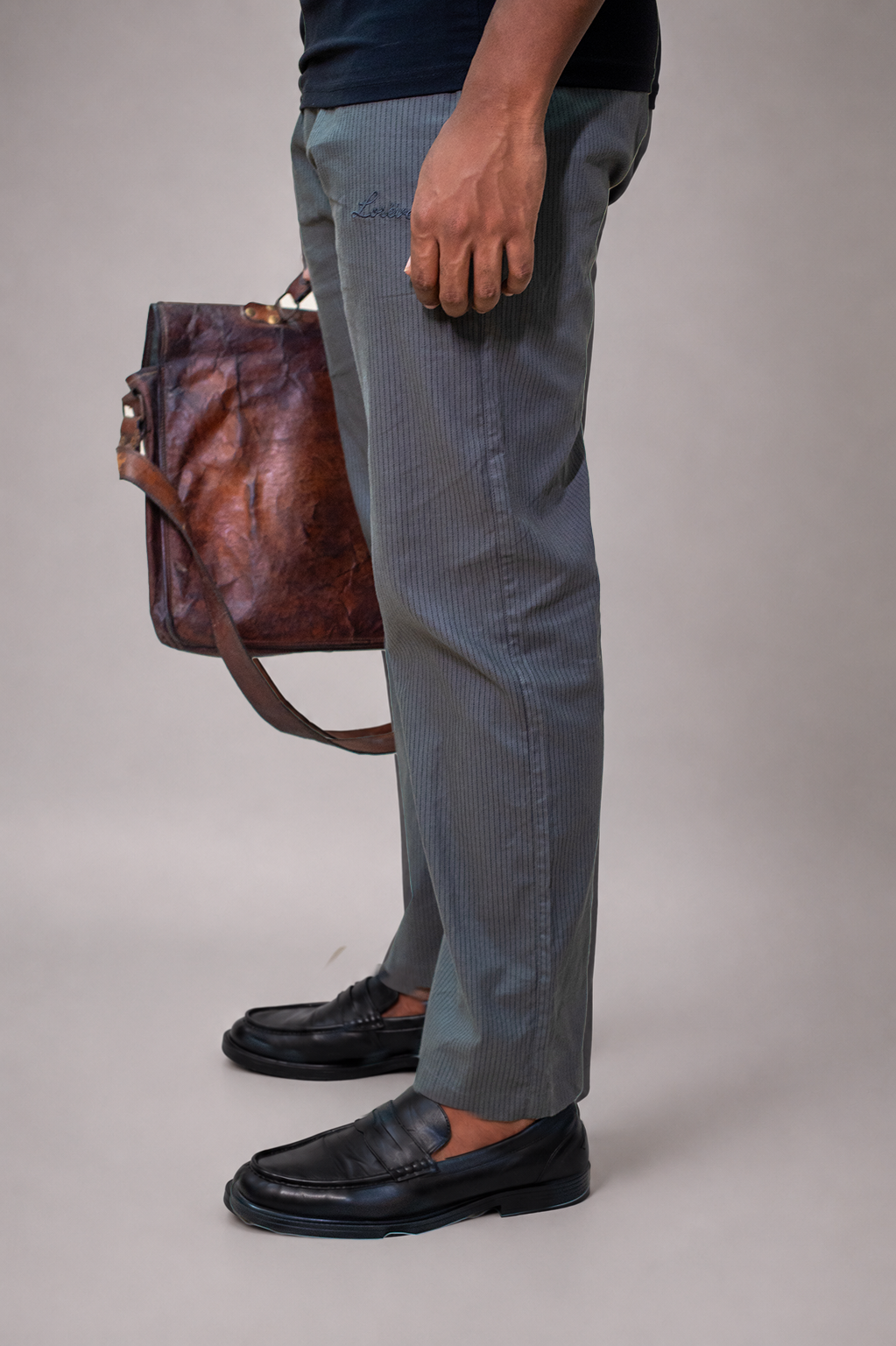 LORËVEN Striped Linen Trouser in Mediterranean Olive — close-up of the trouser leg and black leather loafers with a hand holding a brown leather bag against a grey studio background