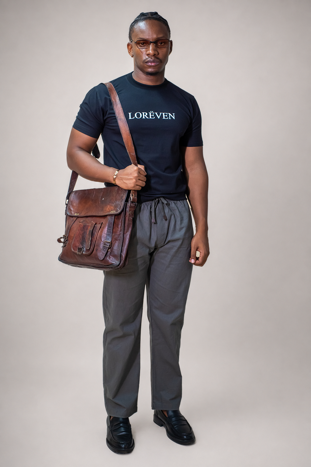 LORËVEN Striped Linen Trouser in Mediterranean Olive — man in a black LORËVEN t-shirt and drawstring trousers carrying a brown leather shoulder bag against a beige background