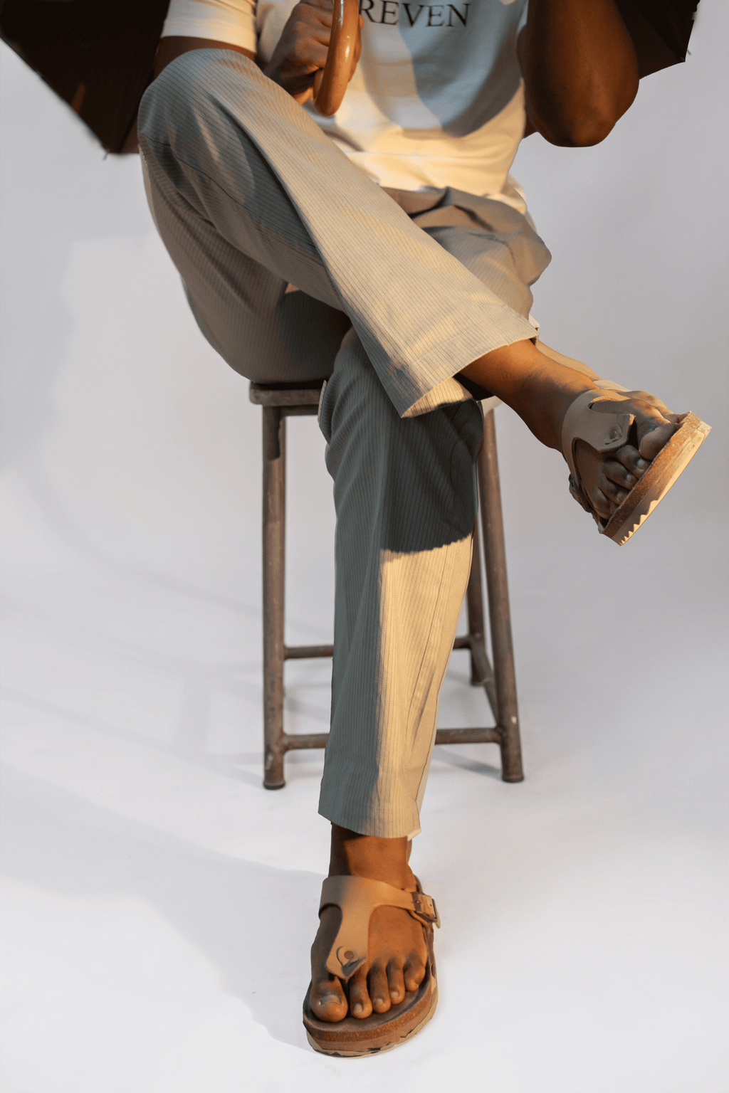 LORËVEN Striped Linen Trouser in Rosemary — close-up of a man seated on a wooden stool wearing wide-leg trousers and brown leather thong sandals against a light grey studio background