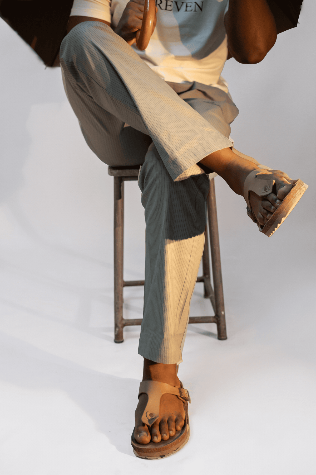 LORËVEN Striped Linen Trouser in Rosemary — close-up of a man seated on a wooden stool wearing wide-leg trousers and brown leather thong sandals against a light grey studio background